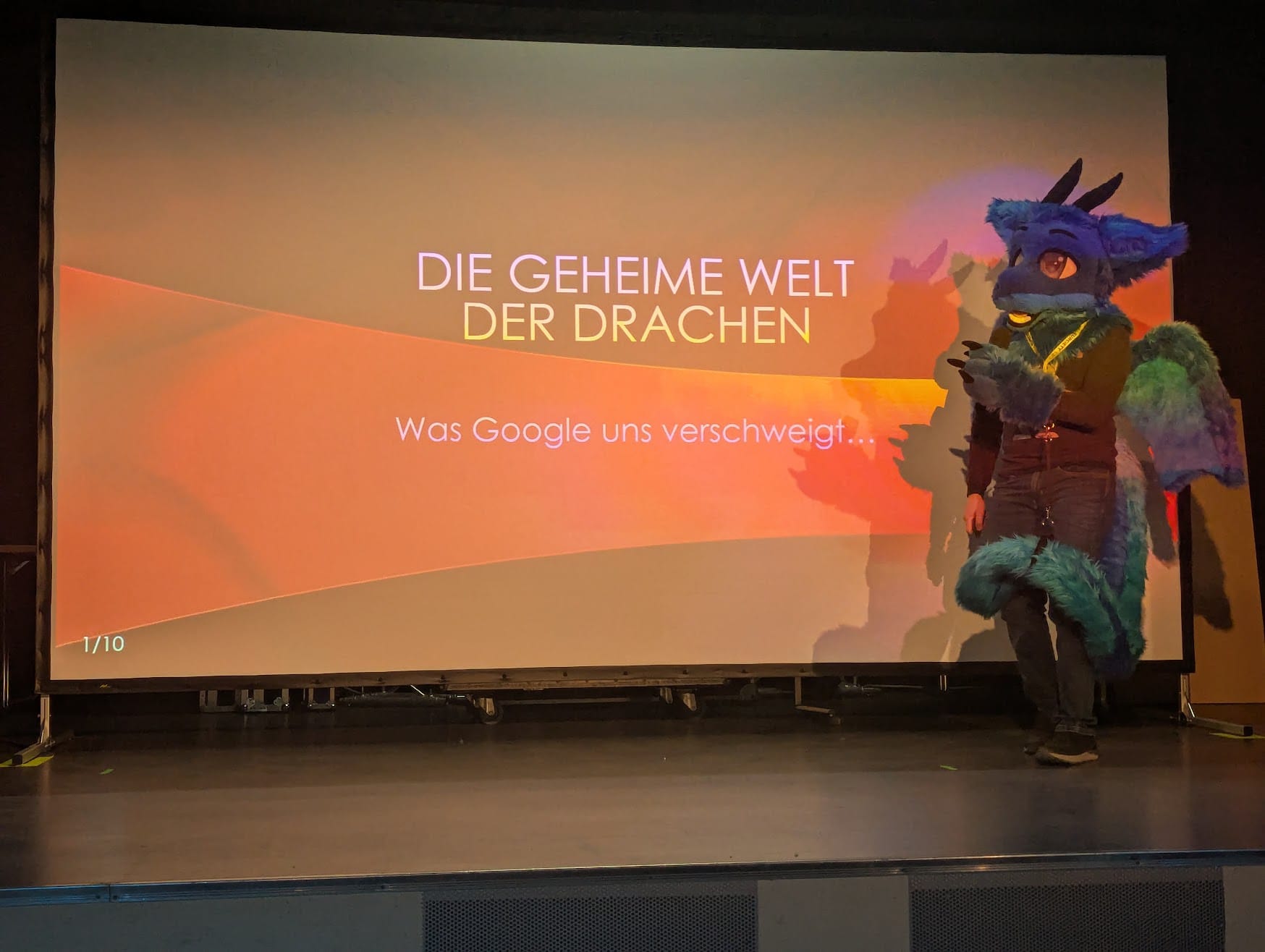 Ein Bild mit mir im Fursuit auf der Bühne, wie ich eine Präsentation über "Die geheime Welt der Drachen" halte.